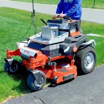 Operator in a blue hoodie driving a red riding lawn mower along a grassy edge beside a paved path.