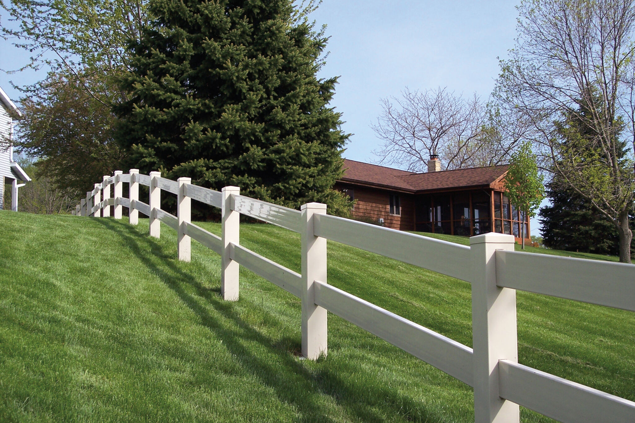 White vinyl post and rail fence along grassy yard near a country side estate.