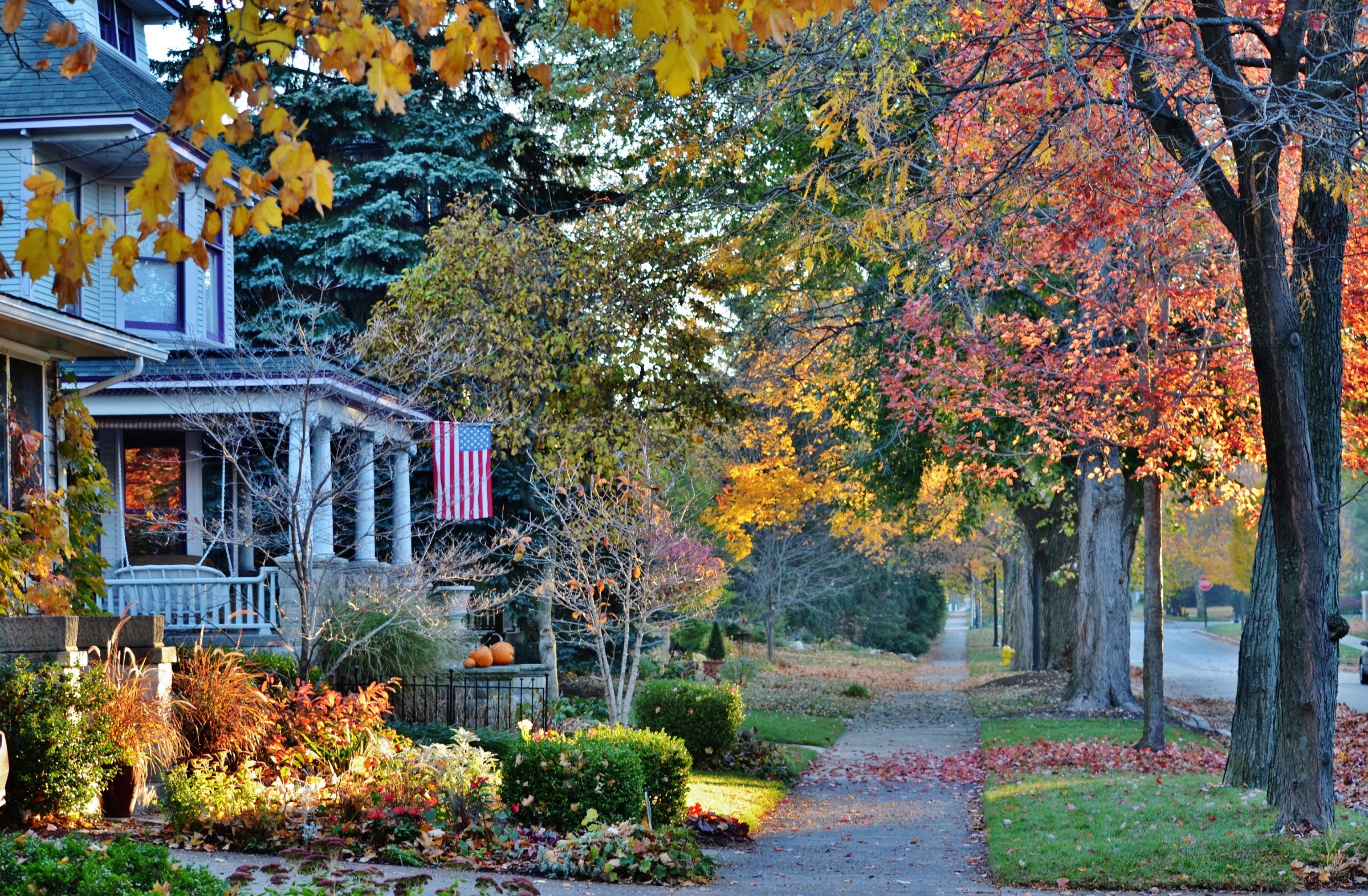 Tree-lined residential street in autumn with a front porch, American flag, and vibrant orange, red, and yellow fall foliage.