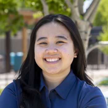 Smiling Asian woman with long dark hair in a blue collared shirt, outdoors on a sunny day.